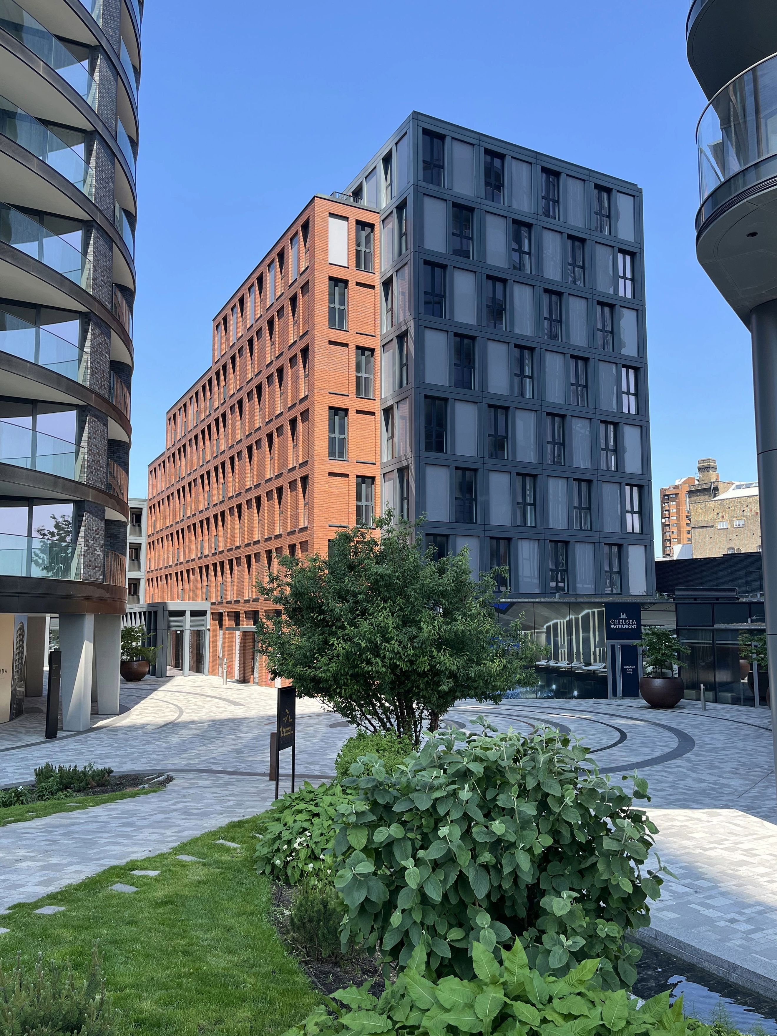 A courtyard surrounded by modern buildings with brick and grey facades, featuring balconies, windows, trees, and landscaping.