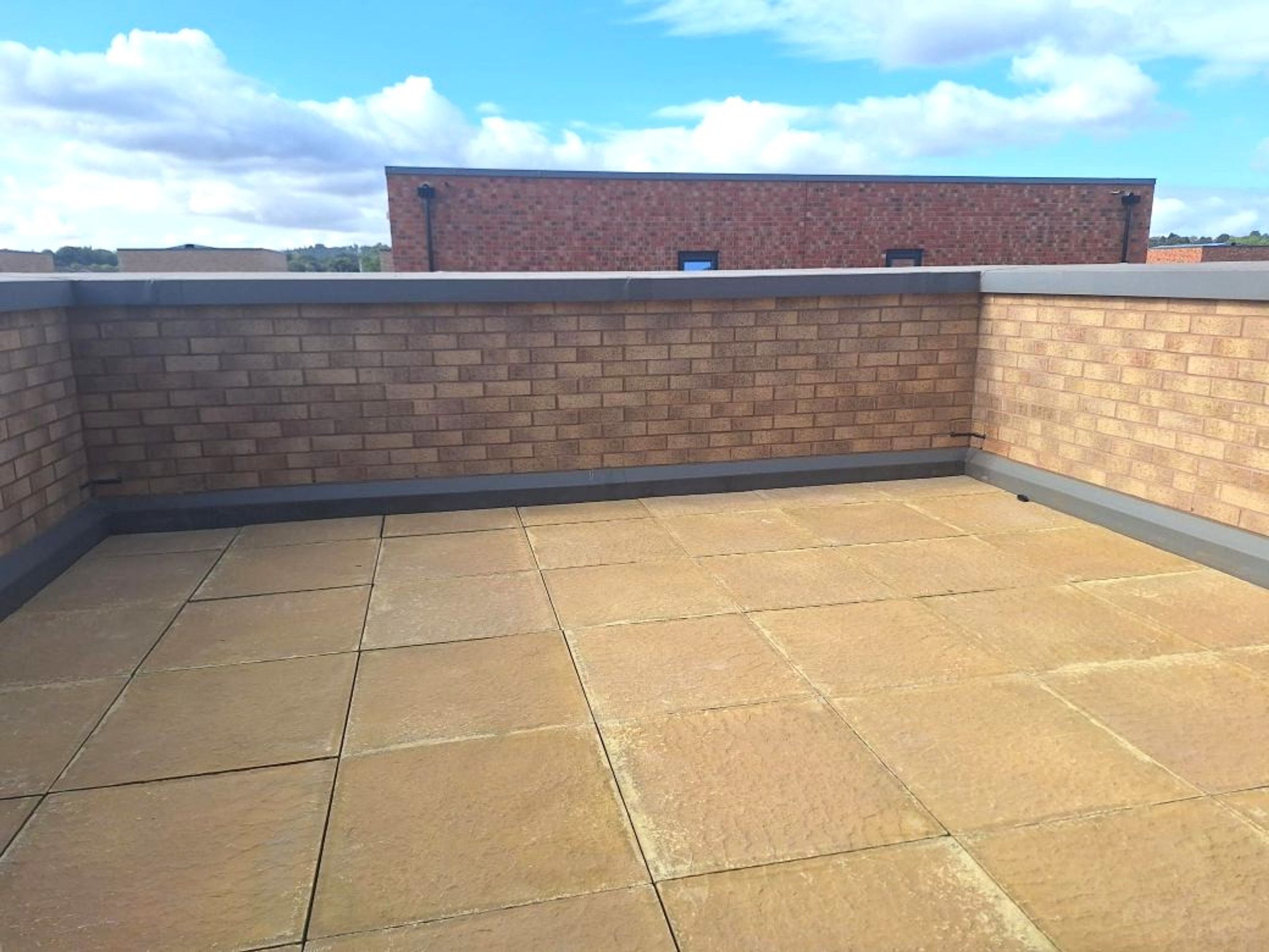 A rooftop terrace with paved flooring and a brick wall under a cloudy blue sky.