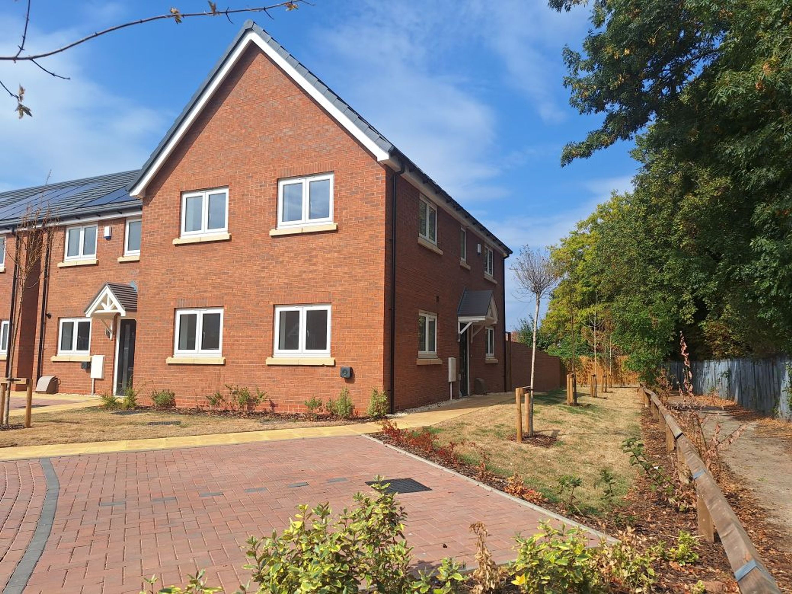 A row of new brick townhouses with white windows and solar panels on the roof, next to a paved driveway and a line of young trees.