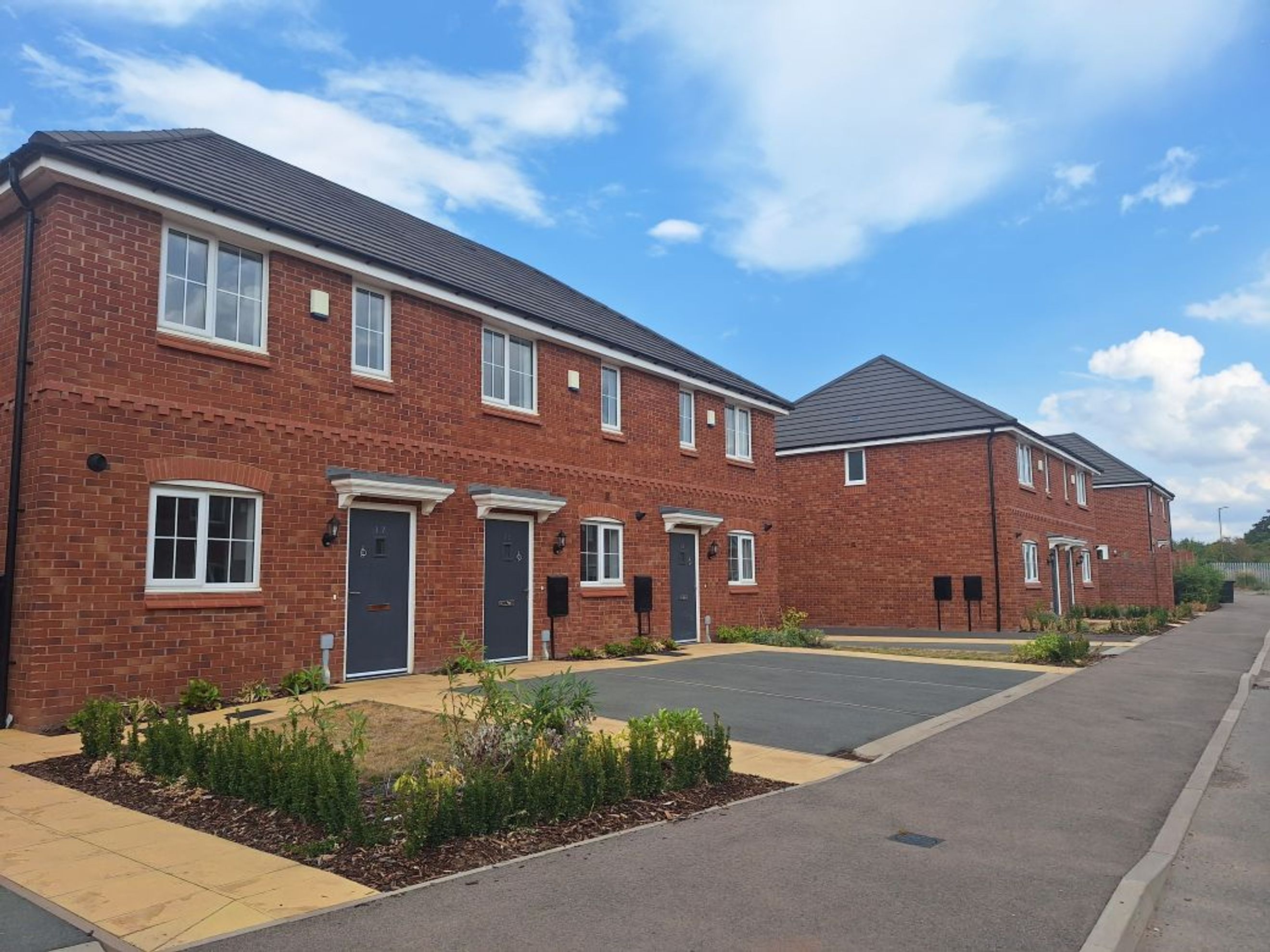 A row of new red brick townhouses with dark grey doors and white windows under a blue sky.