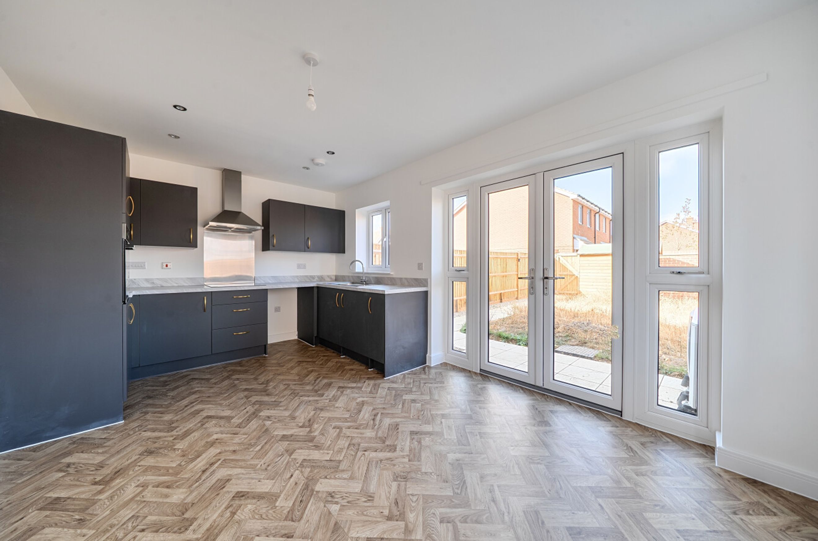 A kitchen with dark cabinets, a stainless steel range hood, and herringbone flooring, opening to a garden through French doors.