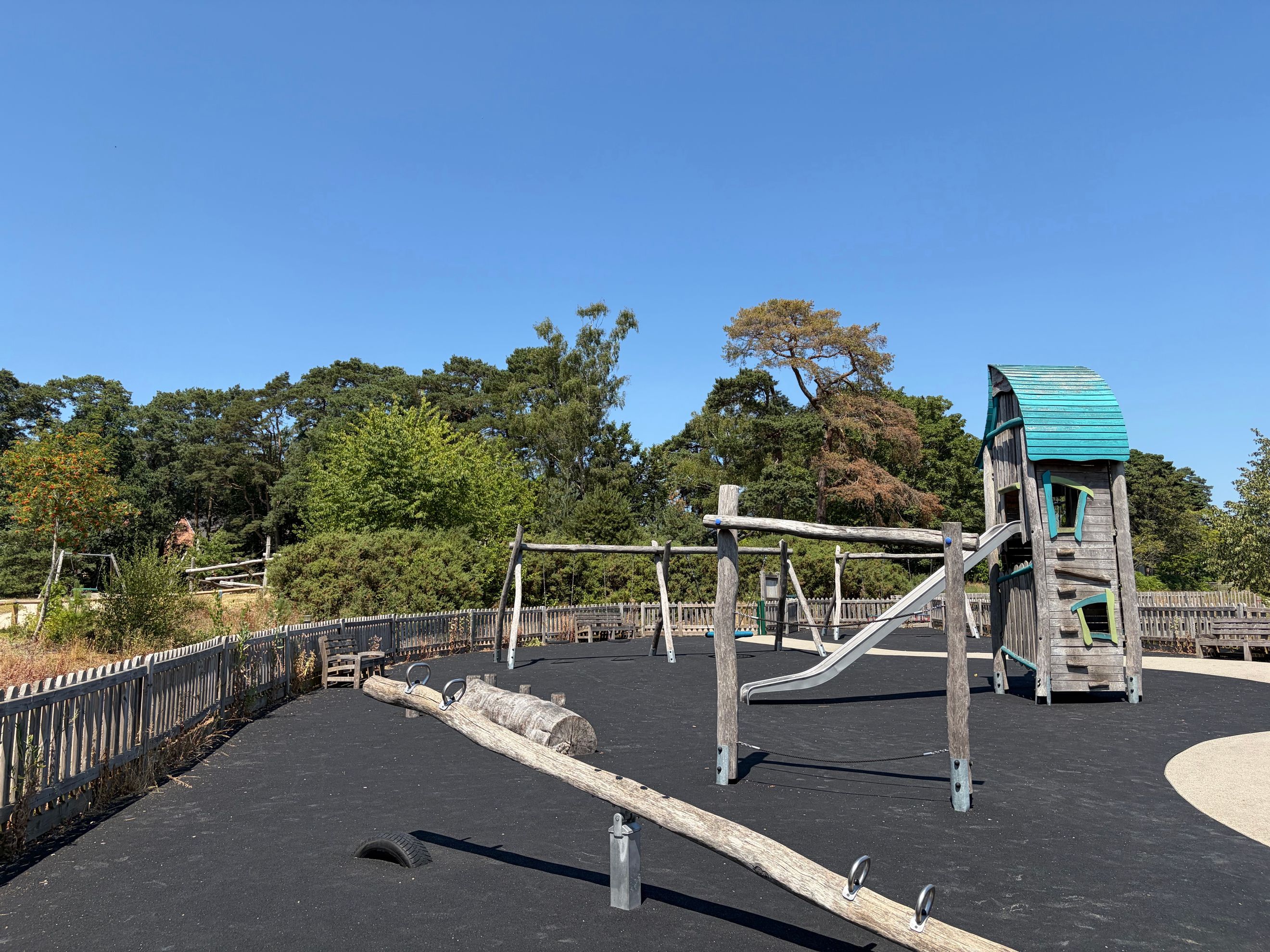A playground with a wooden playhouse, slide, swings, and a seesaw on a rubberized surface.
