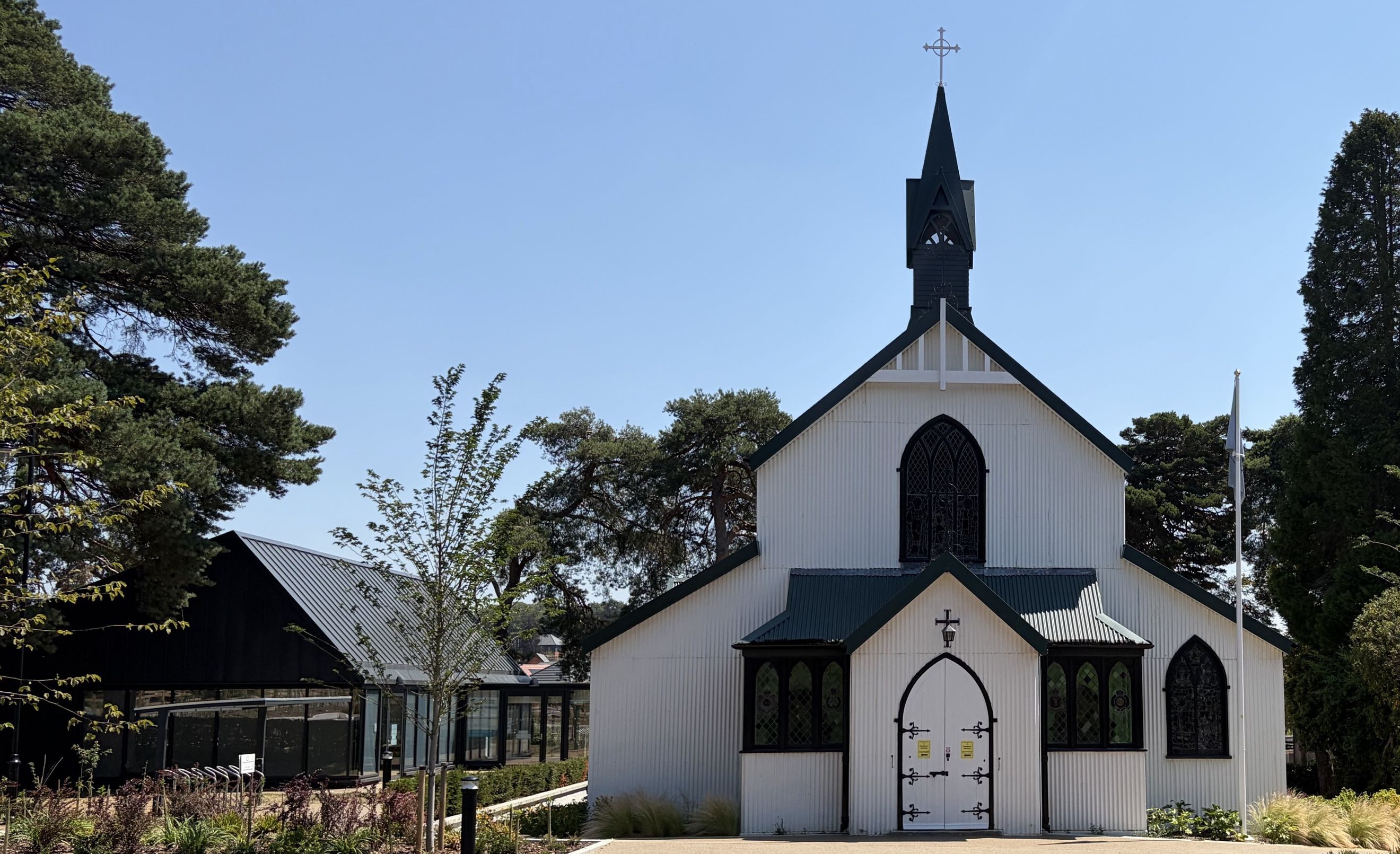 A white corrugated iron church with a dark green roof and steeple, set against a blue sky.
