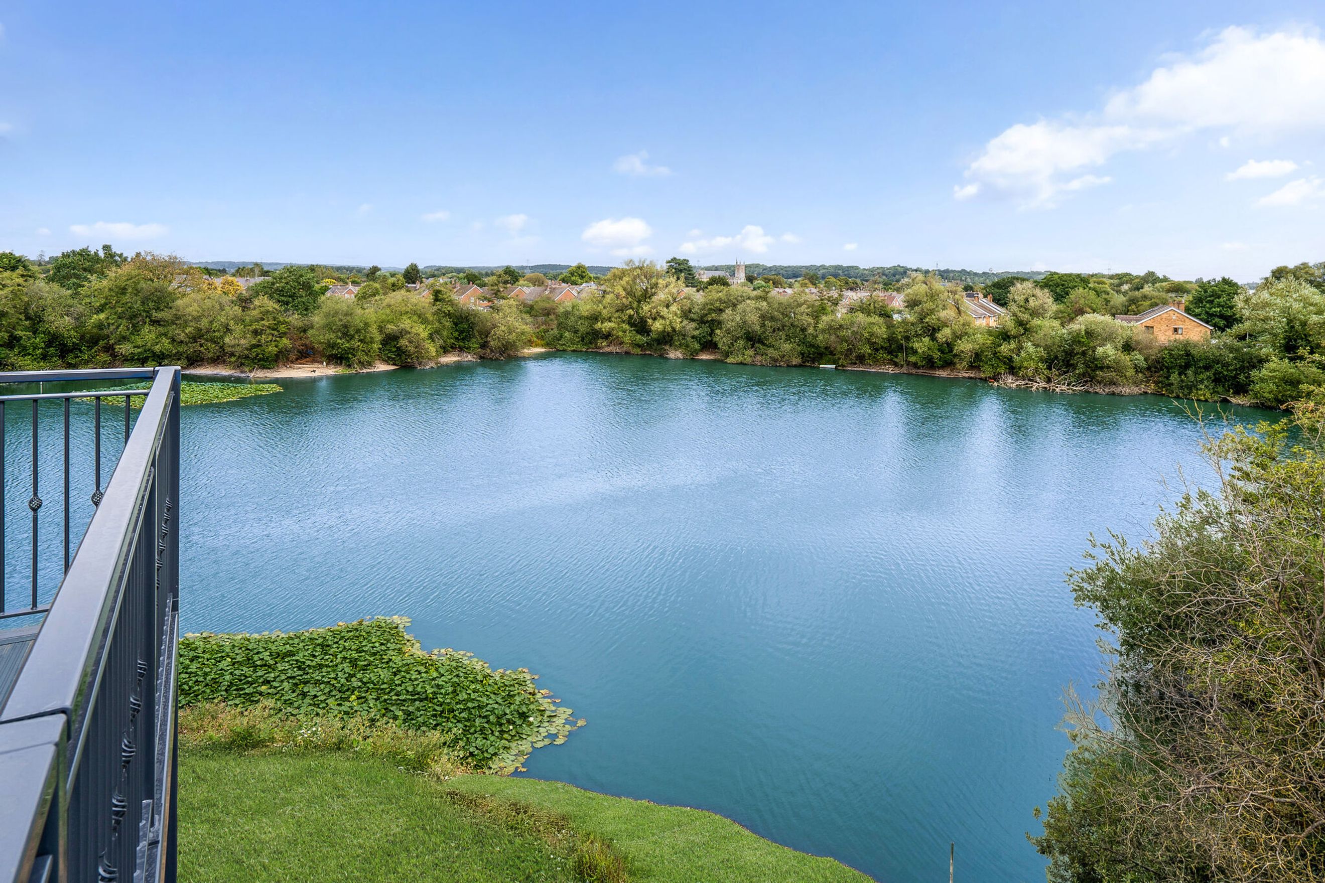 A view of a large blue lake surrounded by trees and houses, with a balcony railing in the foreground.