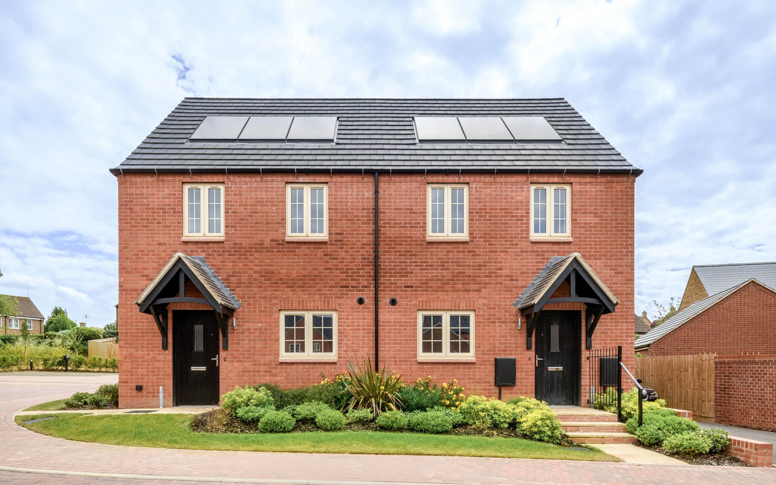 A pair of red brick semi-detached houses with dark doors, cream windows, and solar panels on the roof.
