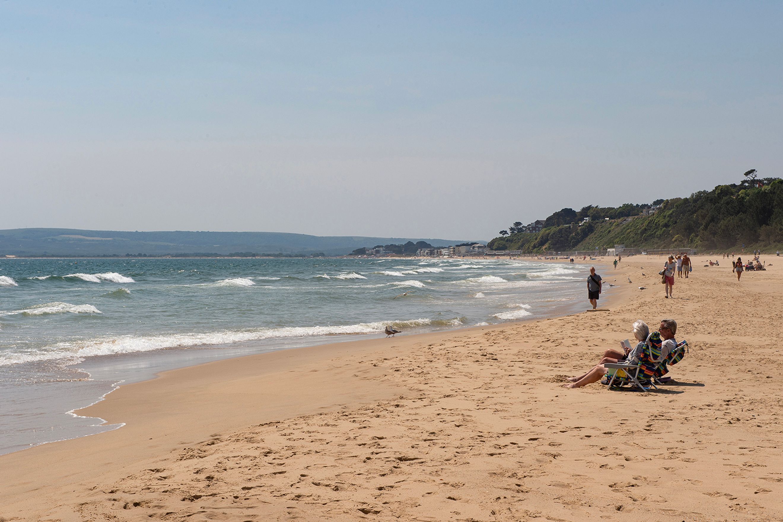 A sandy beach with people relaxing and walking, with the ocean and a tree-covered hill in the background.