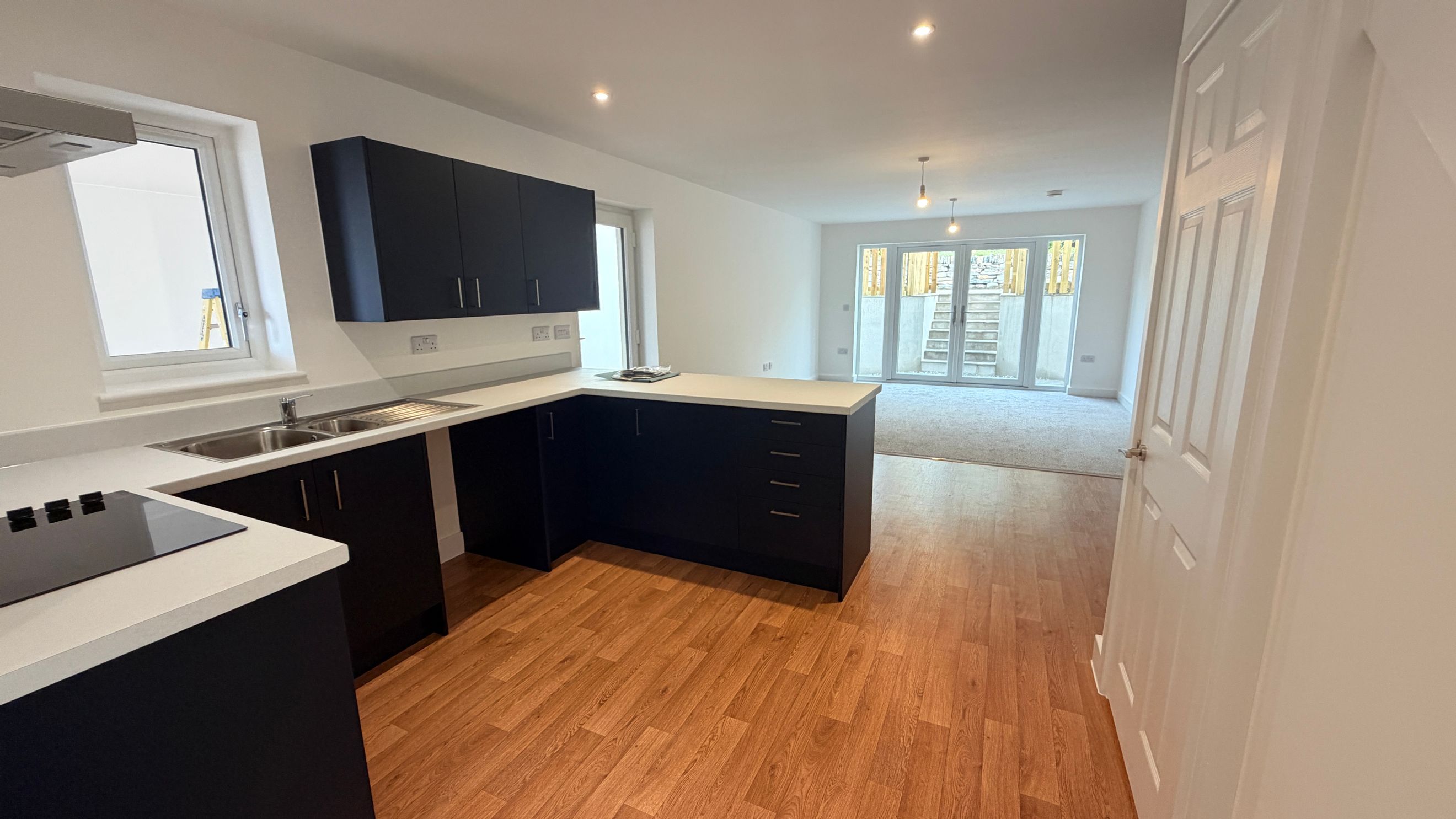 A modern kitchen with dark cabinets and a light countertop, adjacent to a living area with double doors leading outside.