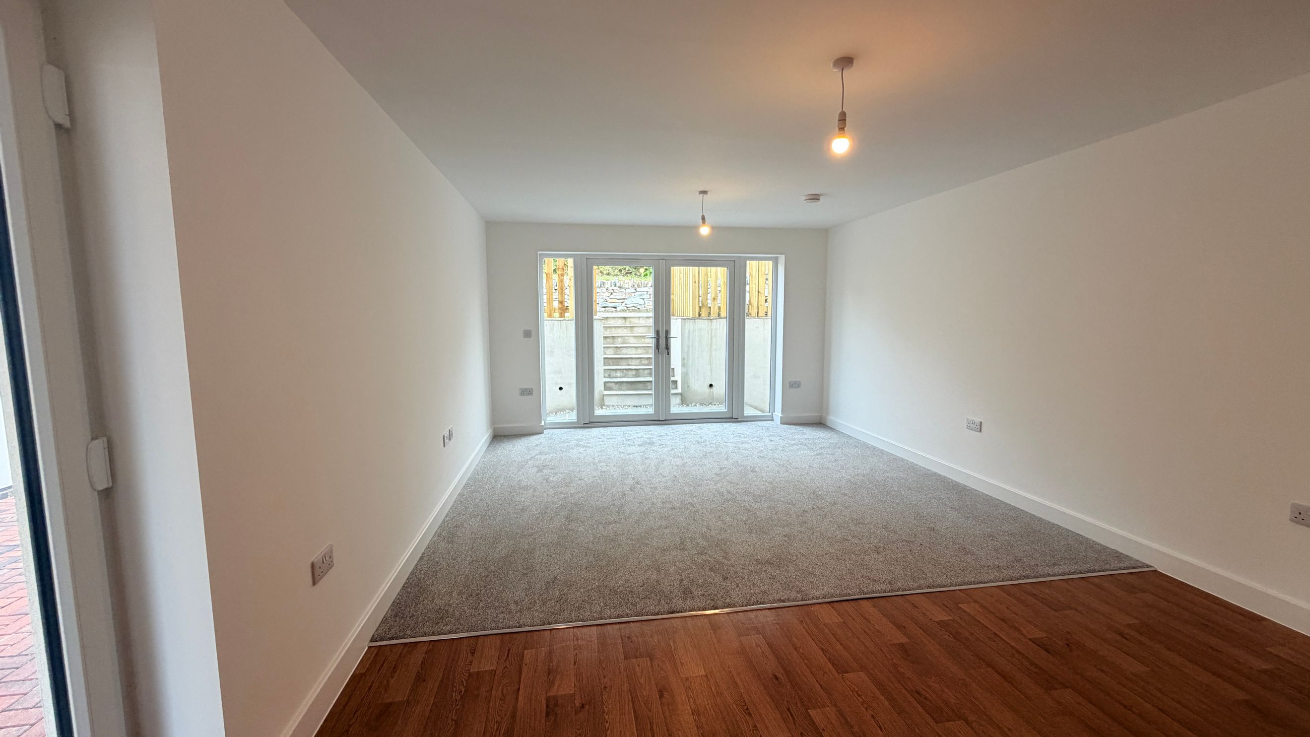 A room with grey carpet and wooden flooring, featuring glass doors leading outside.