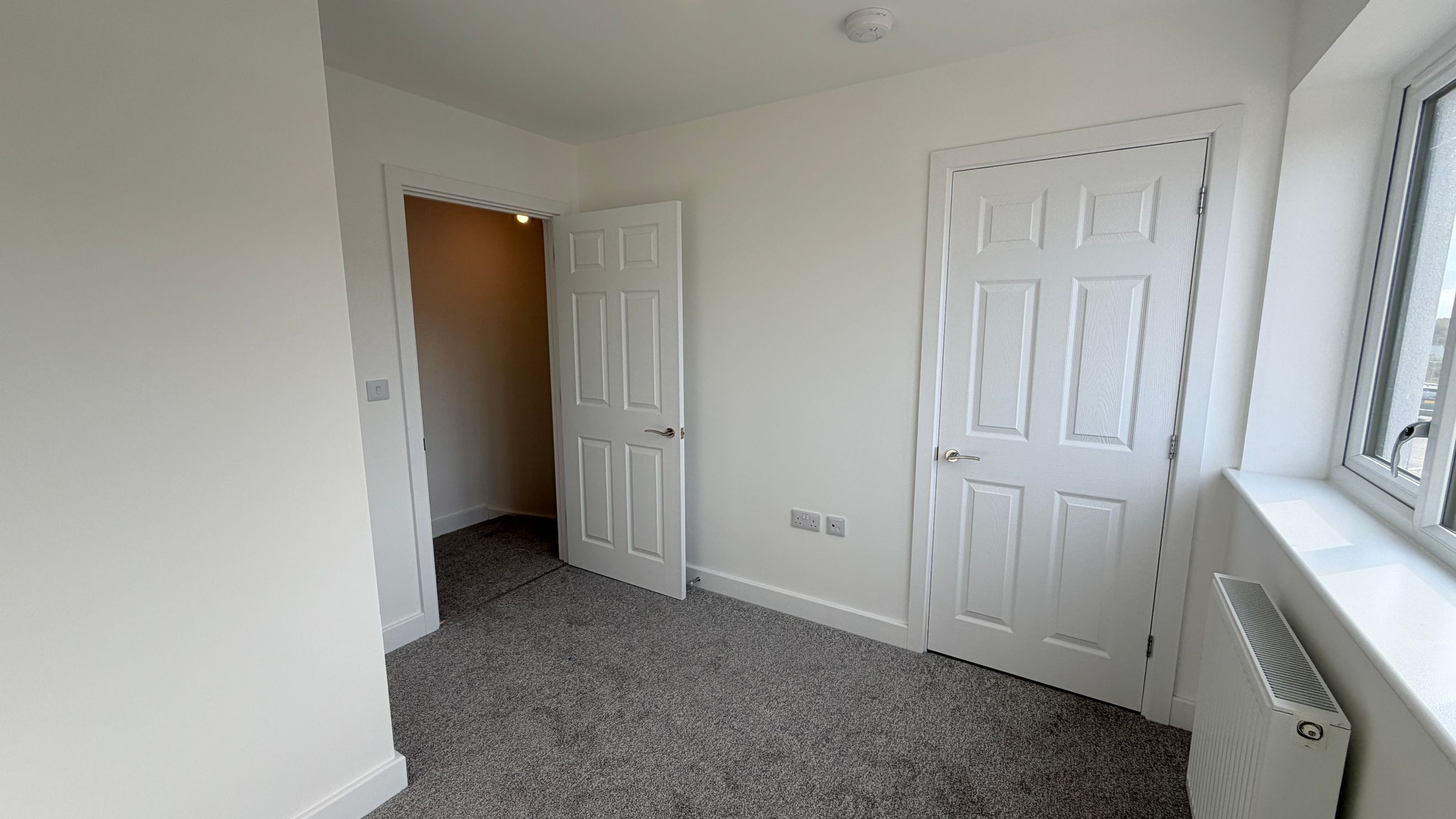 A room with two white doors, a window, a radiator, and grey carpet.