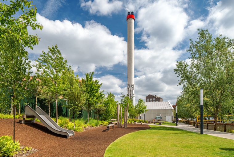 A tall industrial chimney stands in a park with a modern building and trees under a cloudy sky.