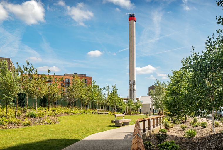 A park with a tall chimney, buildings, trees, and benches.