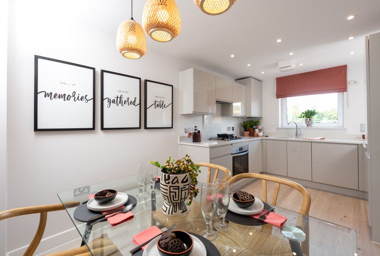 A dining area and kitchen with a glass table, chairs, framed art, pendant lights, and kitchen cabinets.