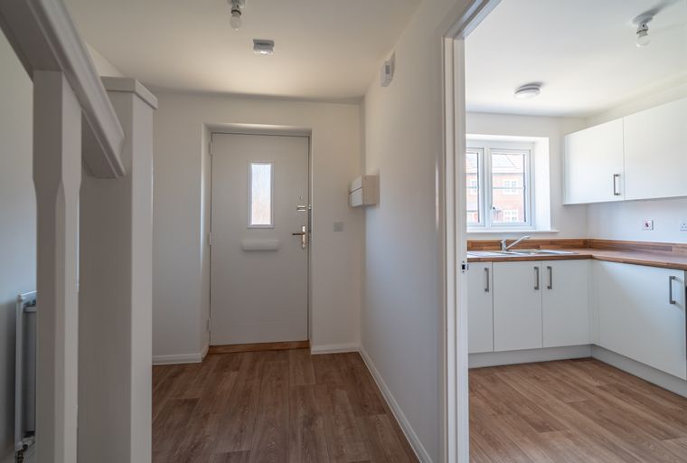 A hallway with a white front door and an adjacent kitchen with white cabinets and a window.