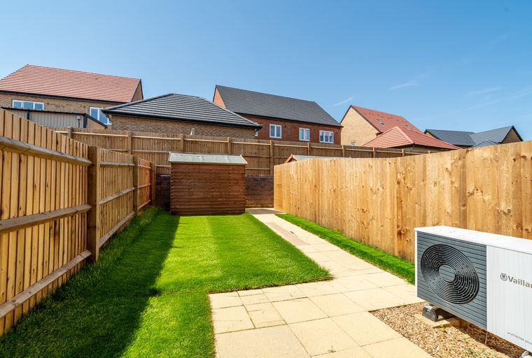 A backyard with a lawn, a shed, a paved path, a heat pump, and a wooden fence.
