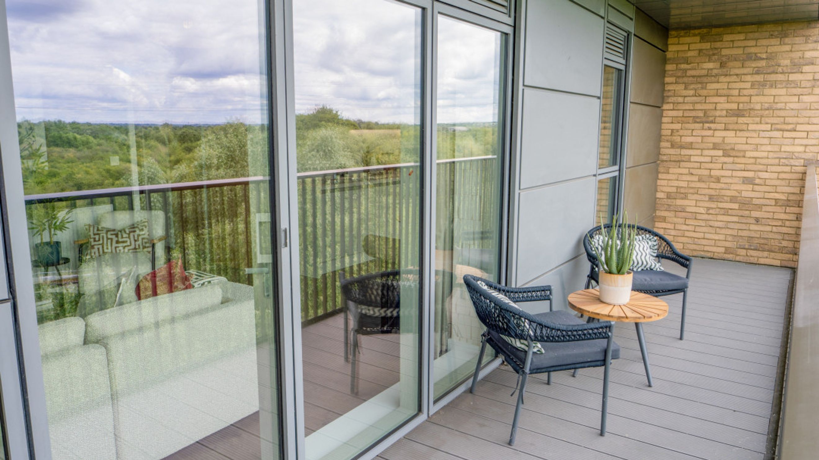 A balcony with two chairs, a table, and a view of trees and sky through glass doors.