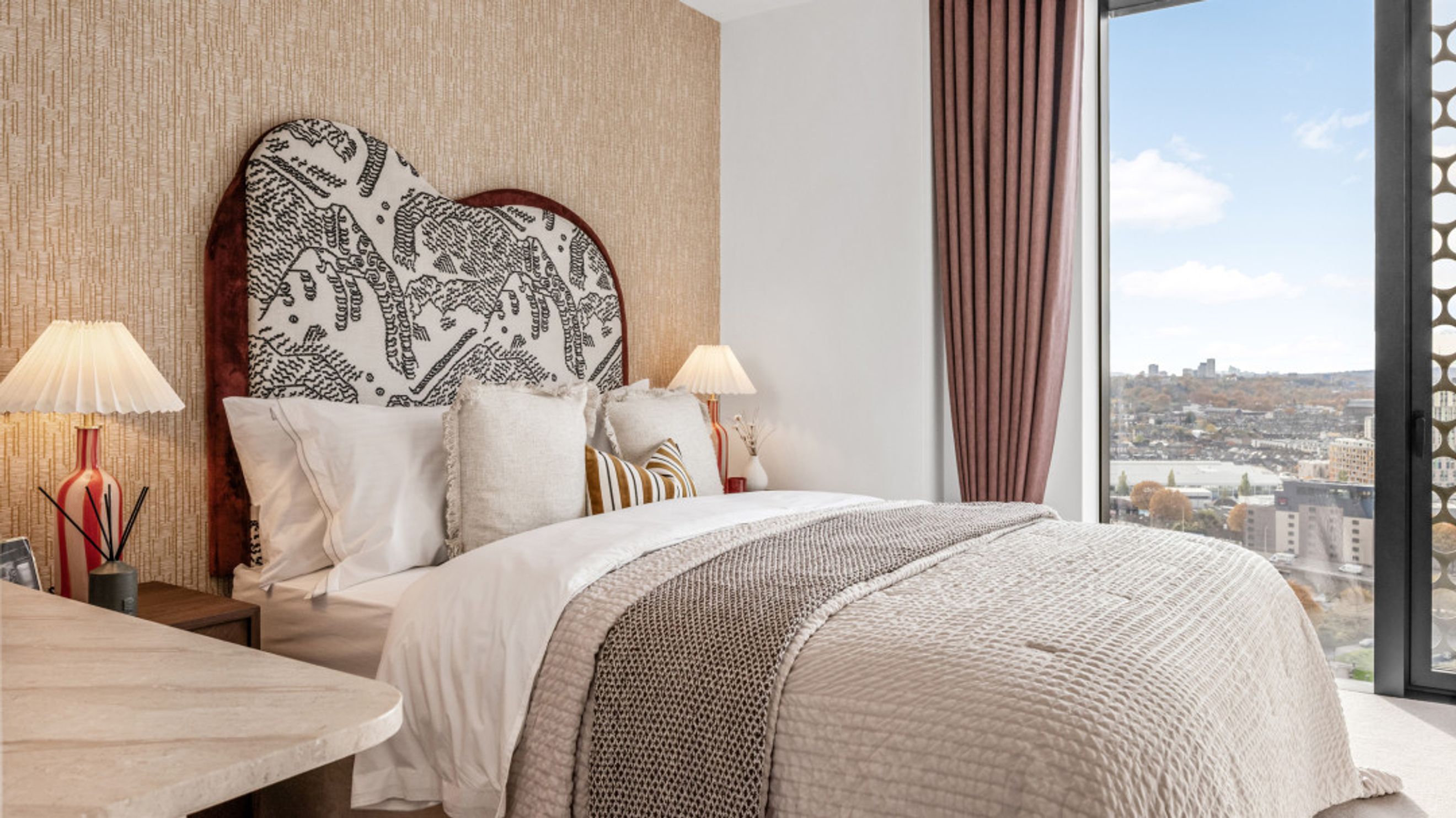 A bedroom with a bed, pillows, lamps, and a large window overlooking a city.