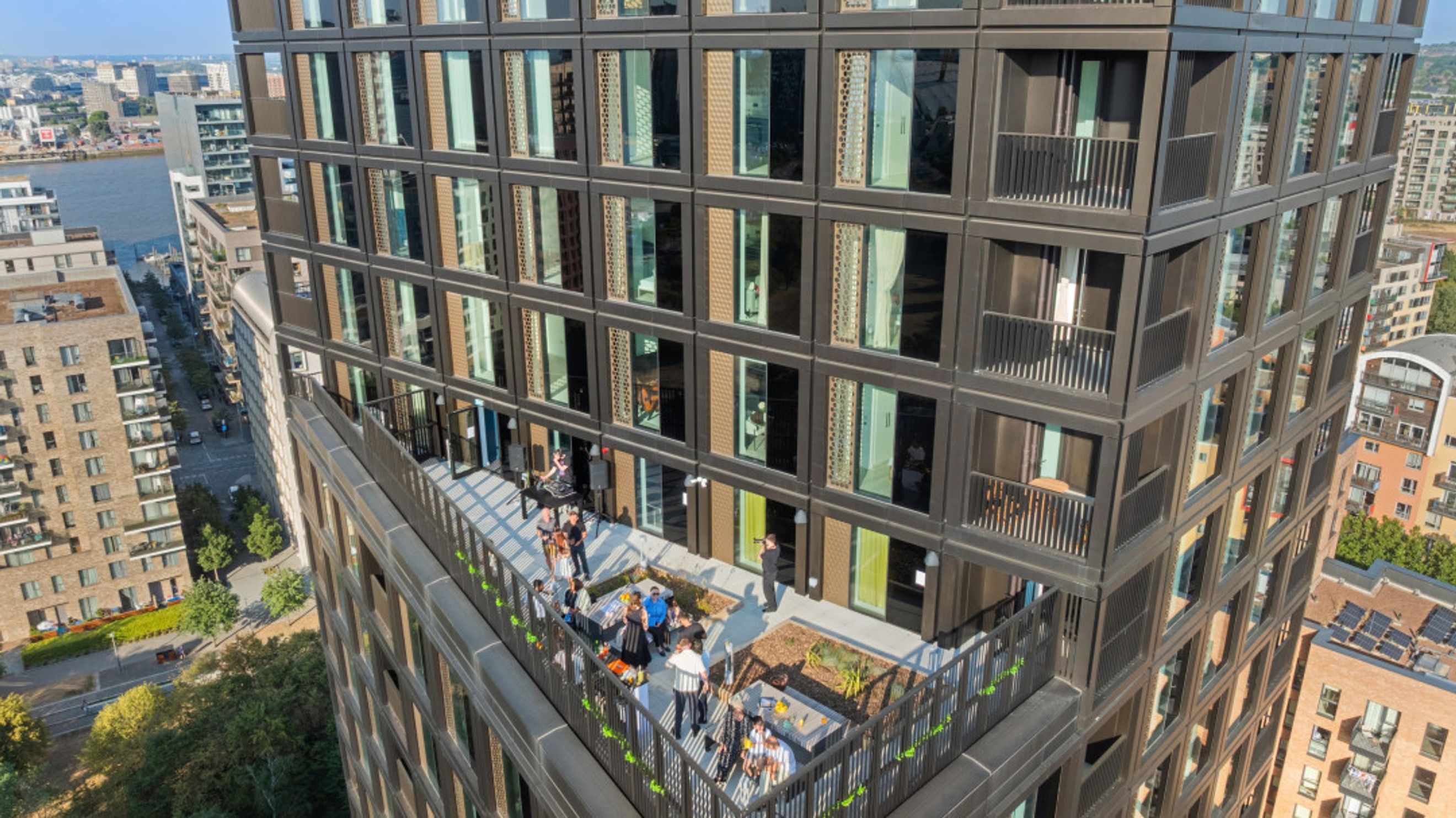 A rooftop terrace with people enjoying a gathering on a sunny day, adjacent to a modern apartment building.