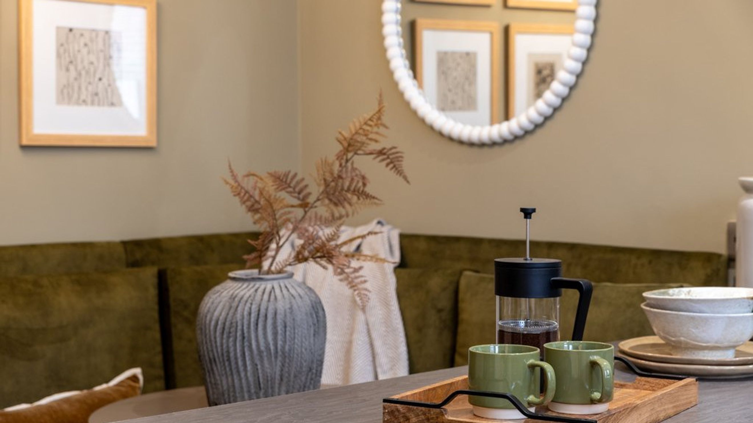 A French press and two mugs sit on a tray in front of a green sofa with a vase of dried flowers.