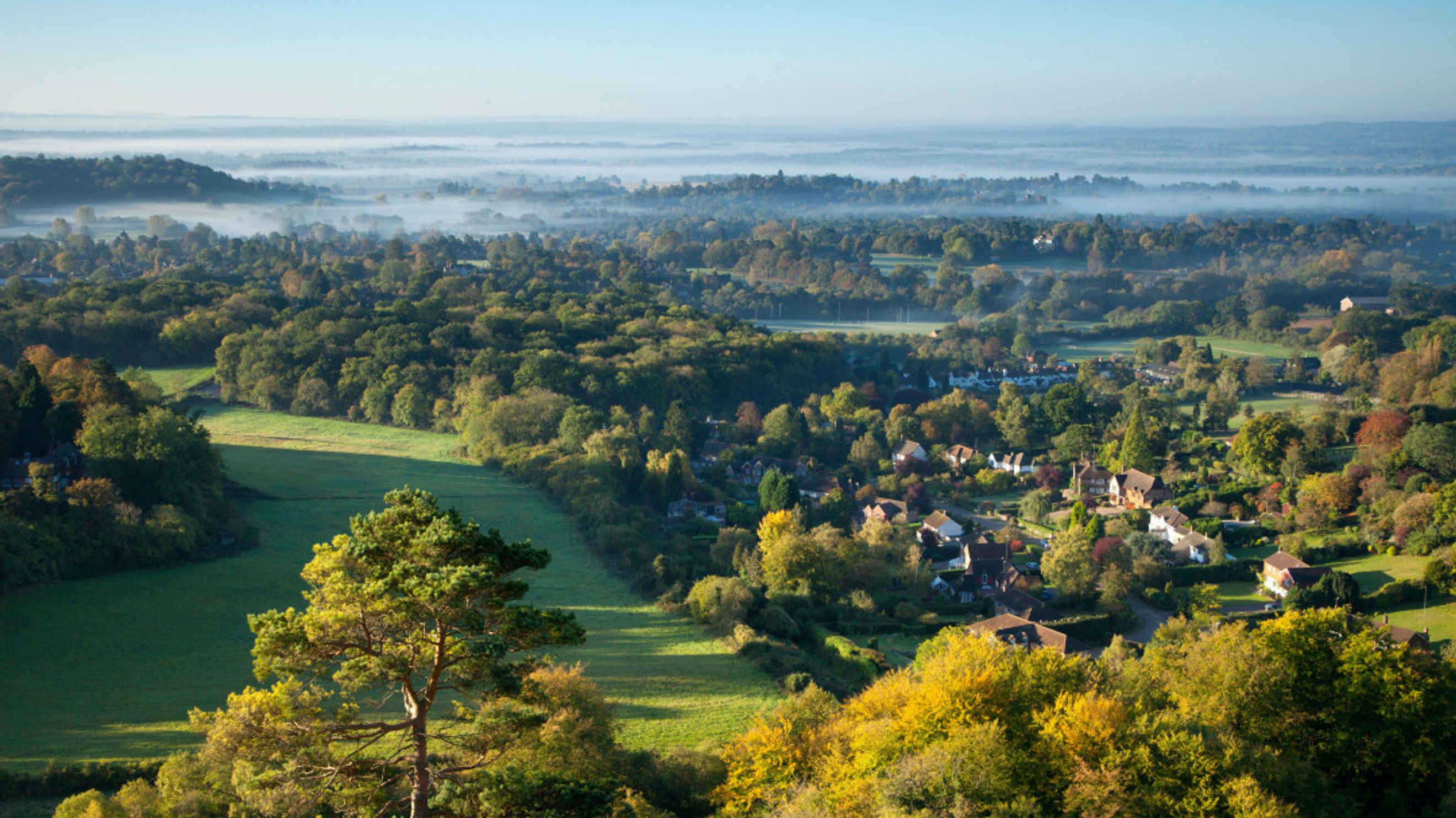 A panoramic view of a village surrounded by trees and hills with mist in the distance.