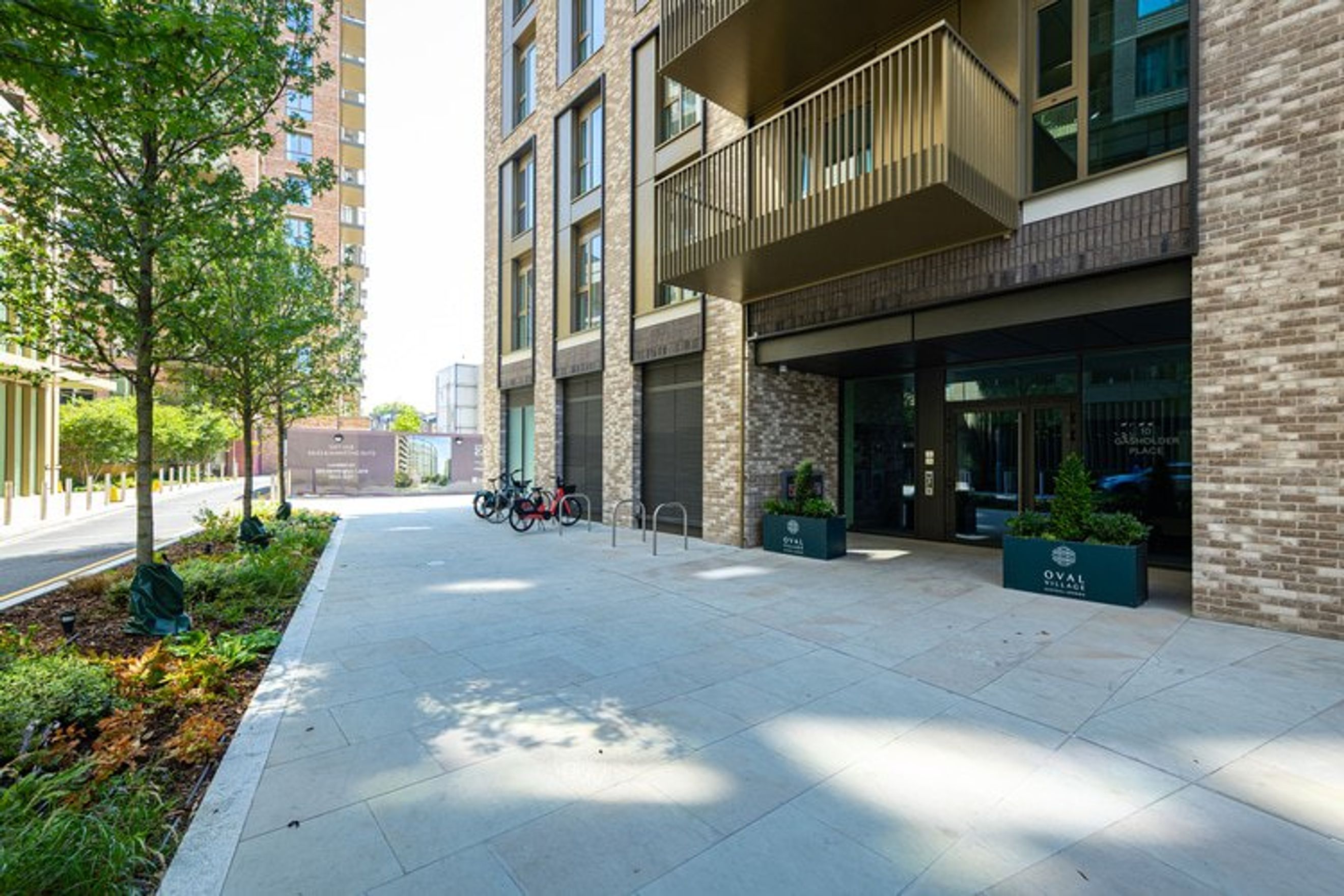 A modern apartment building entrance with bicycles parked outside and planters with the 'Oval Village' logo.