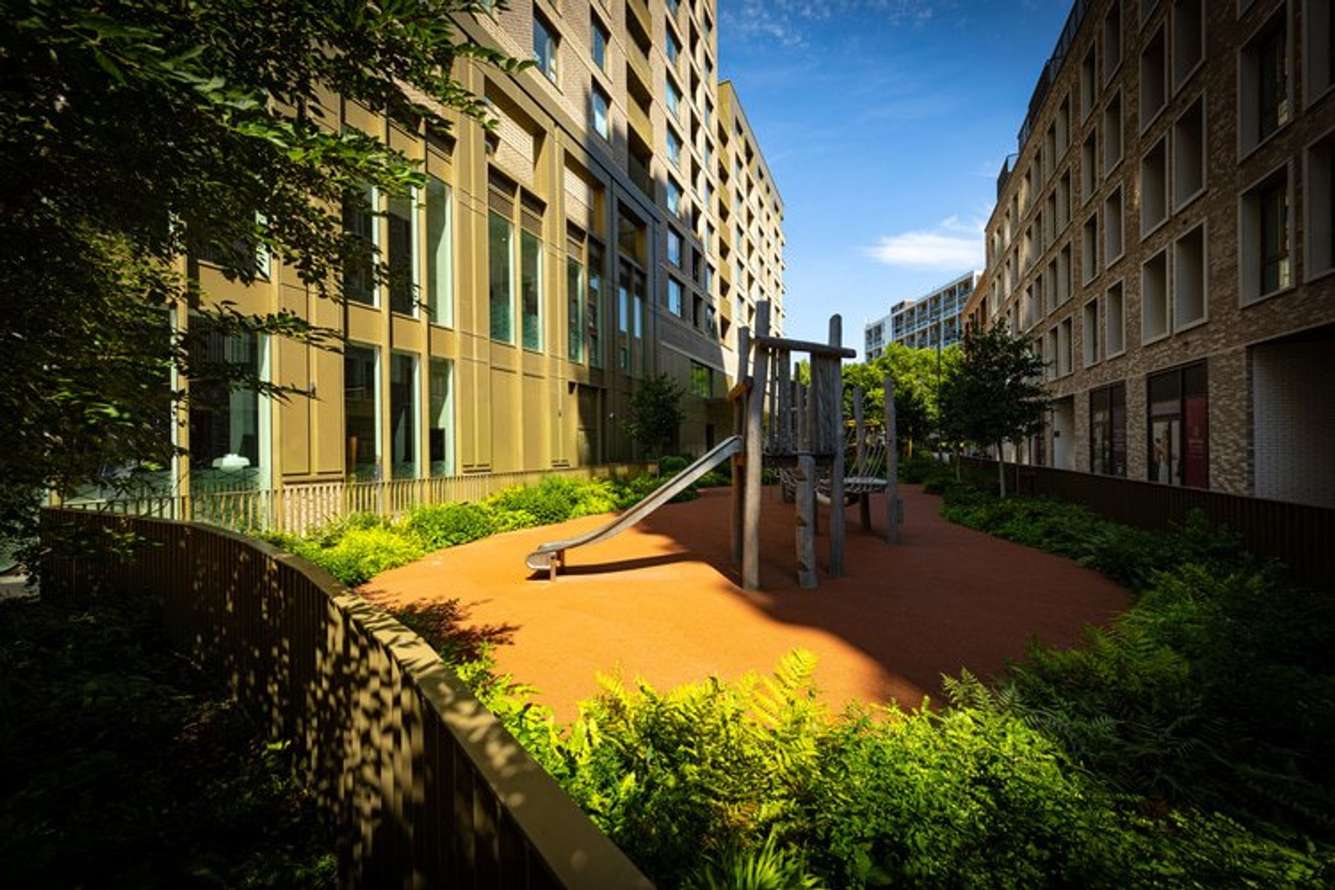 A children's playground with a slide and climbing structure is situated in a courtyard between two modern apartment buildings.