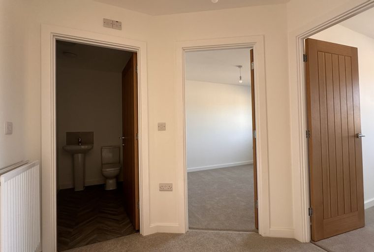 A hallway with three doorways, one leading to a bathroom with a sink and toilet, another to a carpeted room, and a third to a room with a wooden door.