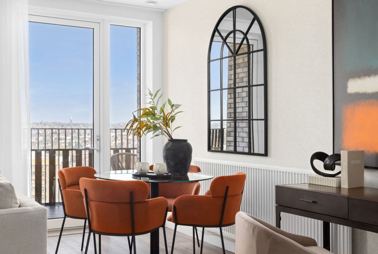 A dining area with a glass table, orange chairs, and a large arched mirror.