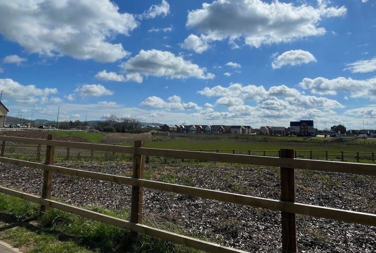 A view of a new housing development with a wooden fence in the foreground and houses in the distance under a cloudy blue sky.