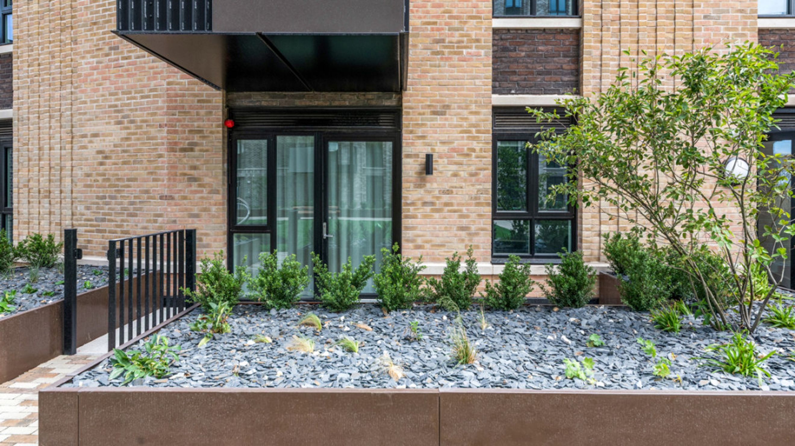 A building exterior with brick walls, large windows, and a landscaped garden with gravel and bushes.