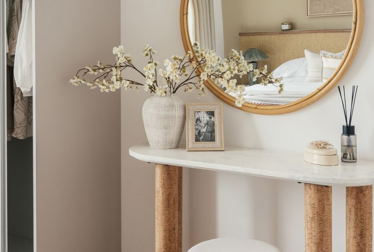 A vanity table with a marble top and cork legs, a mirror, a vase with flowers, and a reed diffuser.