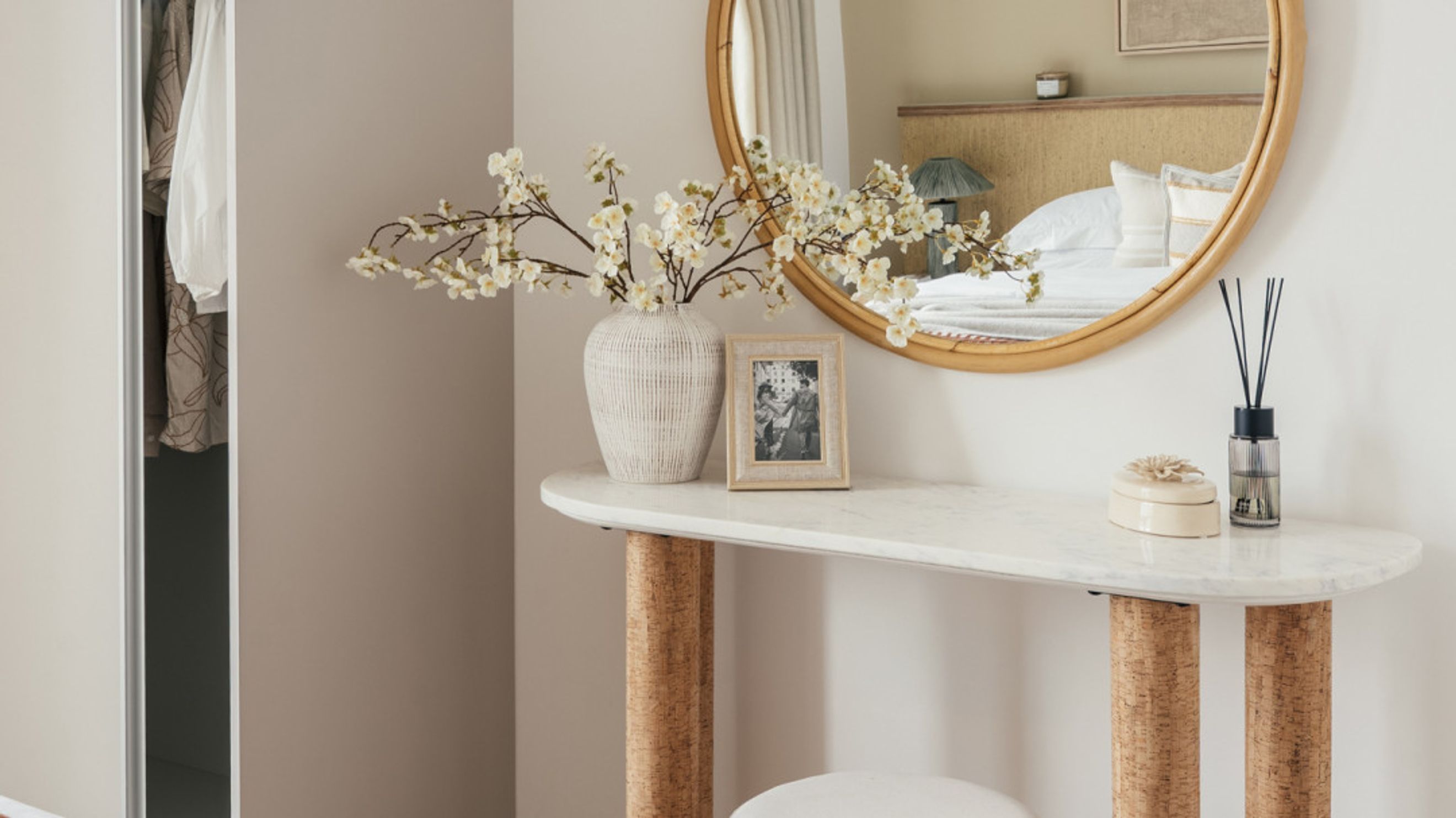 A vanity table with a marble top and cork legs, a mirror, a vase with flowers, and a reed diffuser.