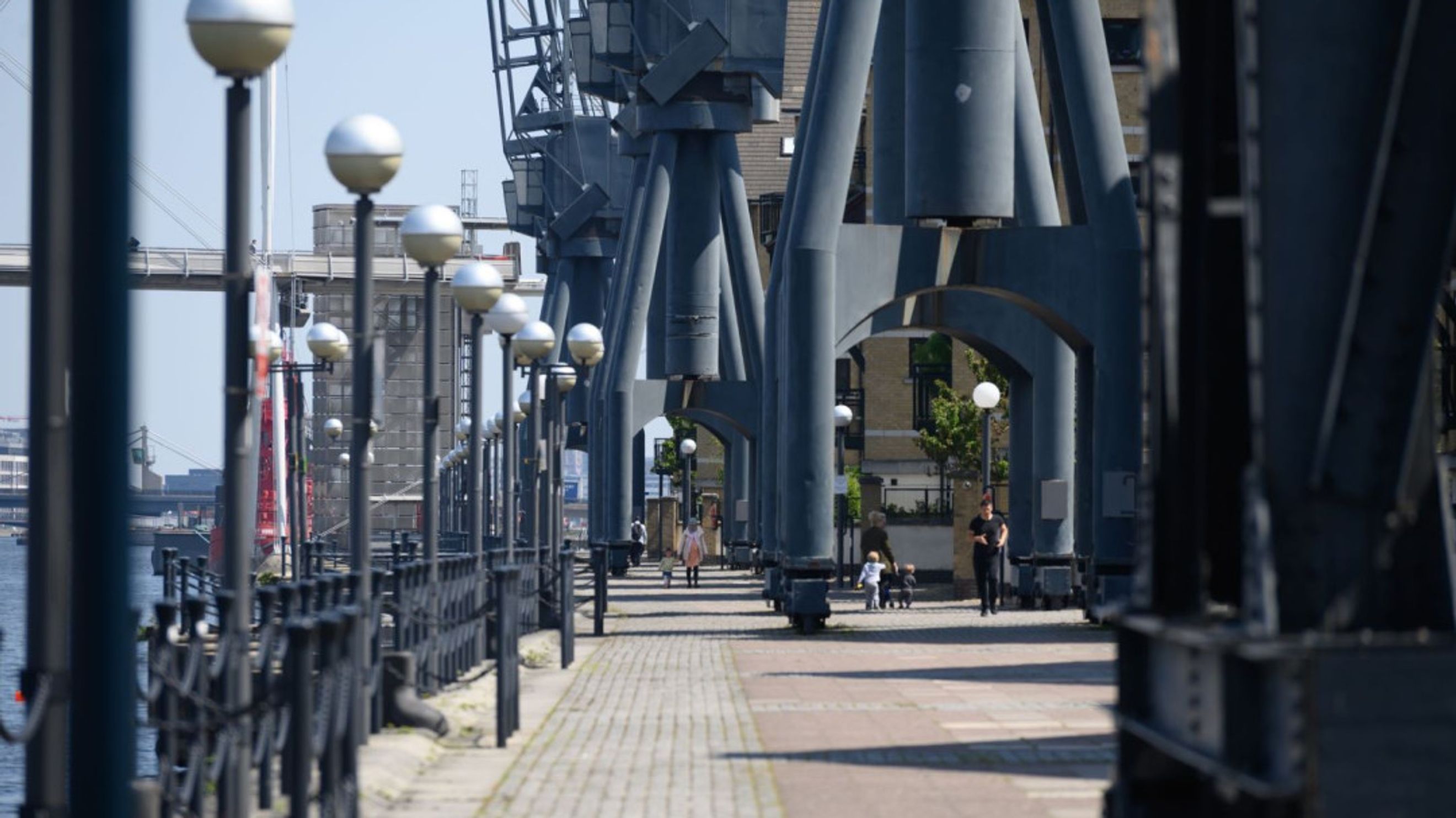 A paved walkway lined with lampposts and large industrial cranes along a body of water.