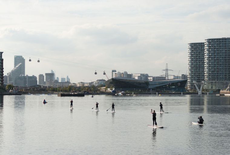 A city skyline with modern buildings and cable cars over a body of water with people paddleboarding.