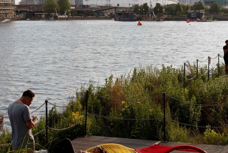 A man stands by a river while another person relaxes on a hammock.