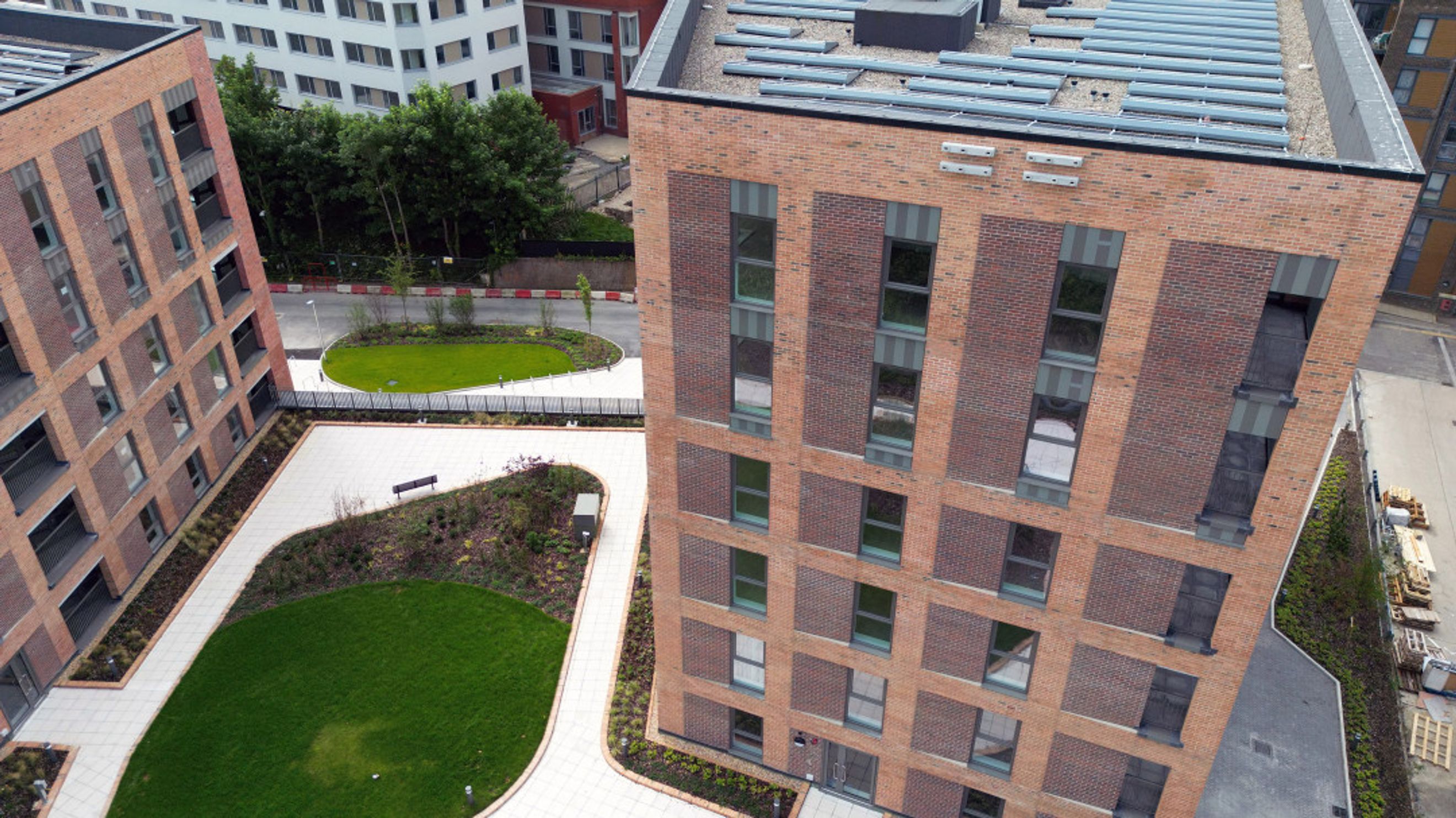 An aerial view of a modern brick apartment building with balconies and solar panels on the roof, surrounded by green spaces and paved walkways.