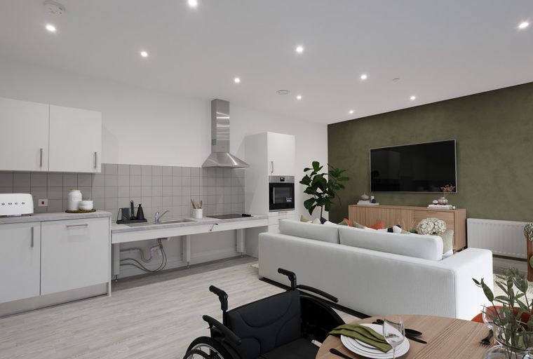 A modern open-plan kitchen and living area with a sofa, TV, and a wheelchair.