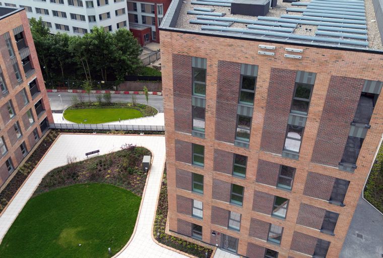 An aerial view of a modern brick apartment building with balconies and solar panels on the roof, surrounded by green spaces and paved walkways.