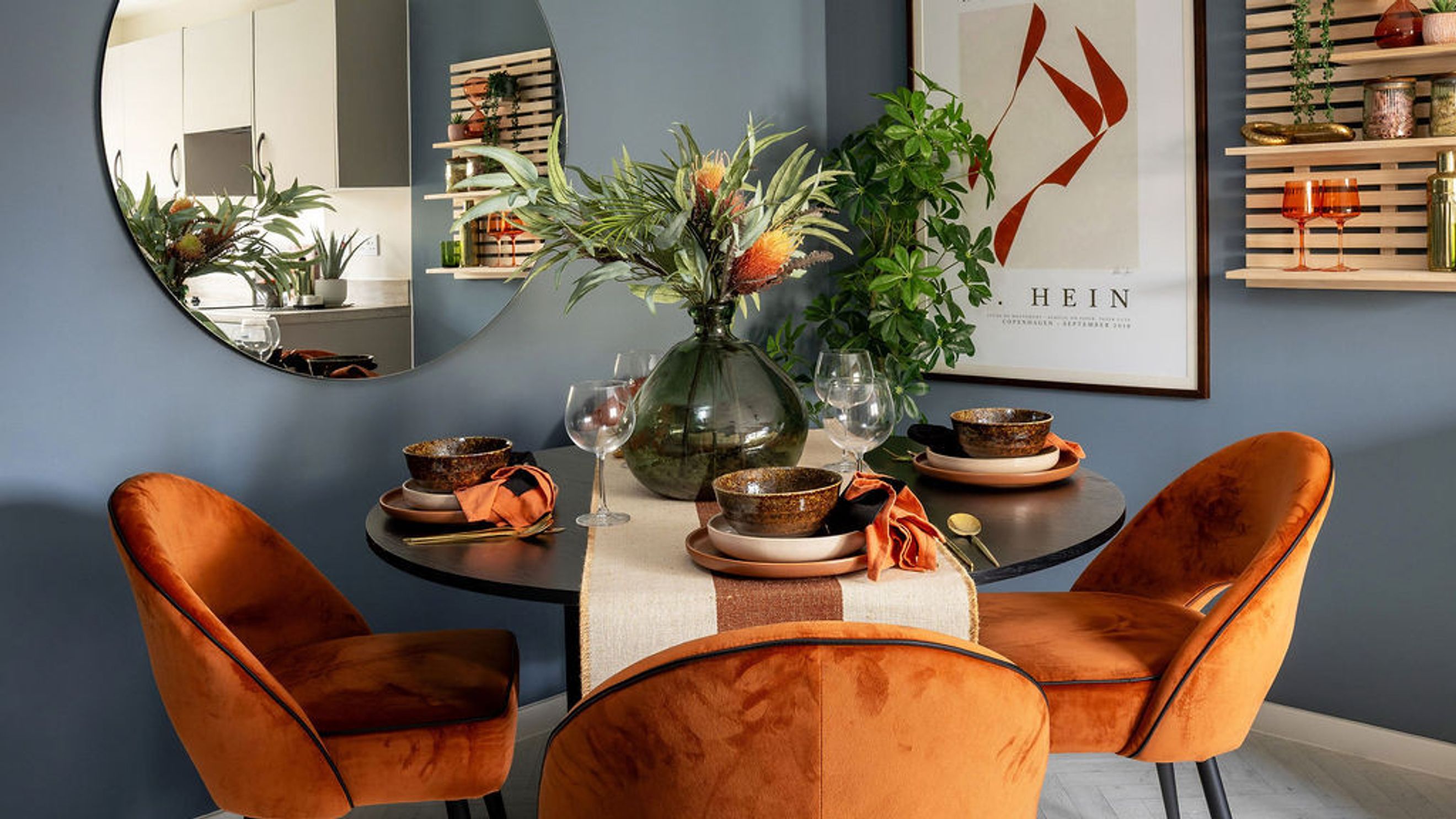 A dining table set for two with orange velvet chairs and a floral centerpiece.