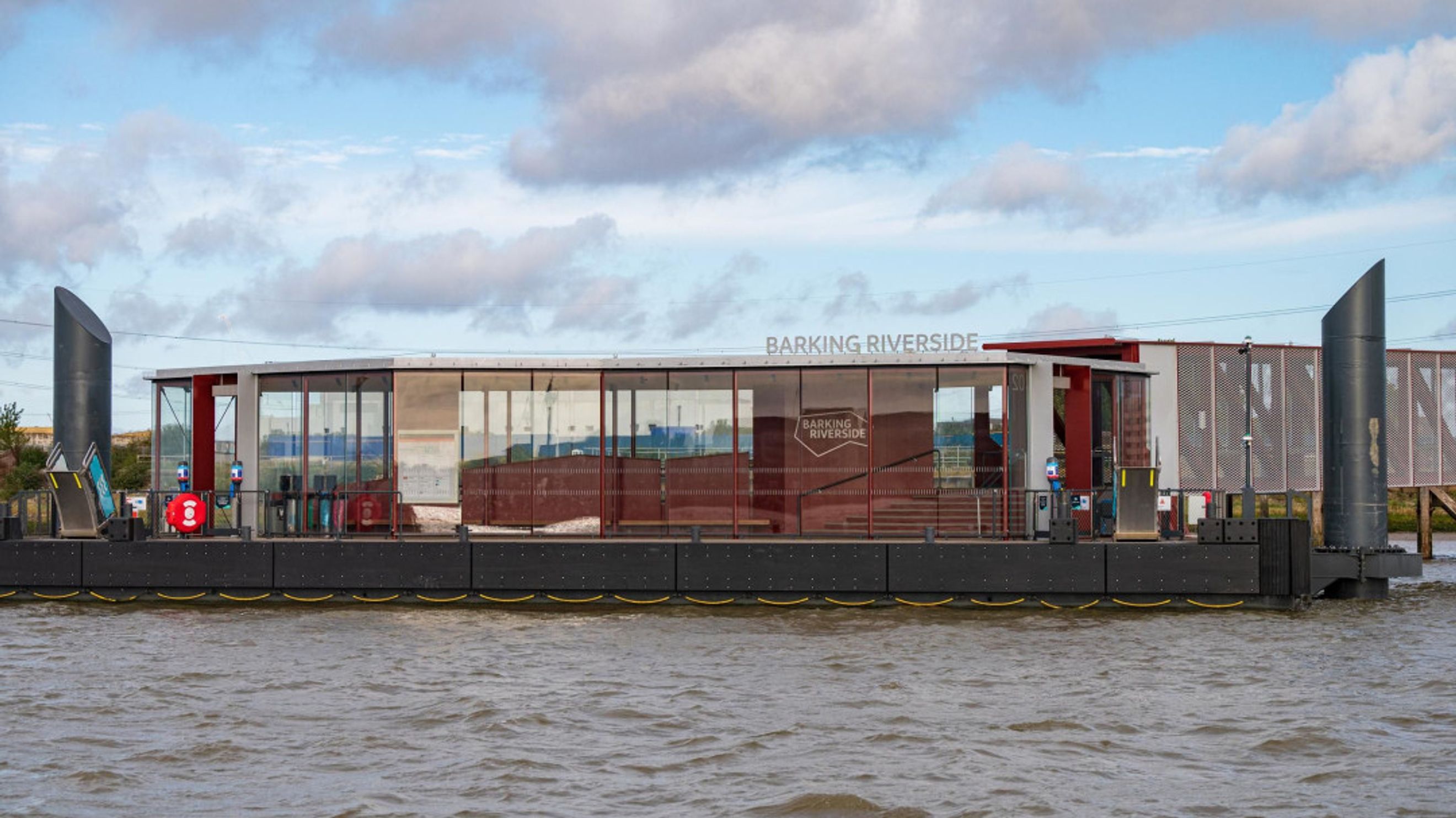 A modern pier structure with glass walls and 'BARKING RIVERSIDE' signage on the water.