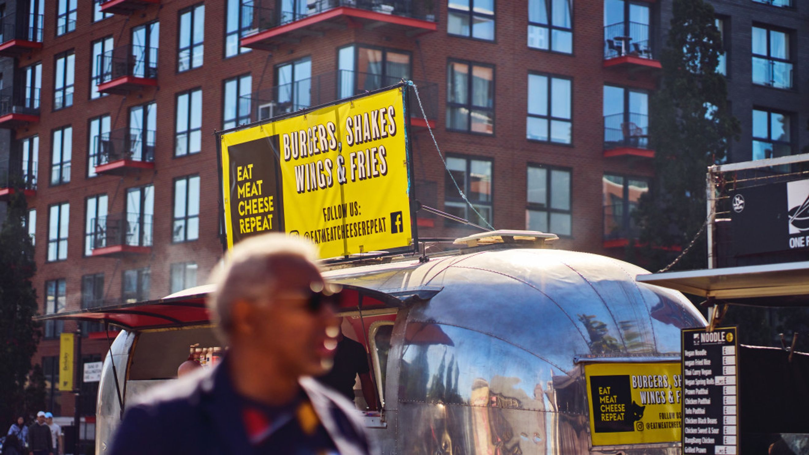A food truck with a sign advertising burgers, shakes, wings, and fries in front of a modern apartment building.