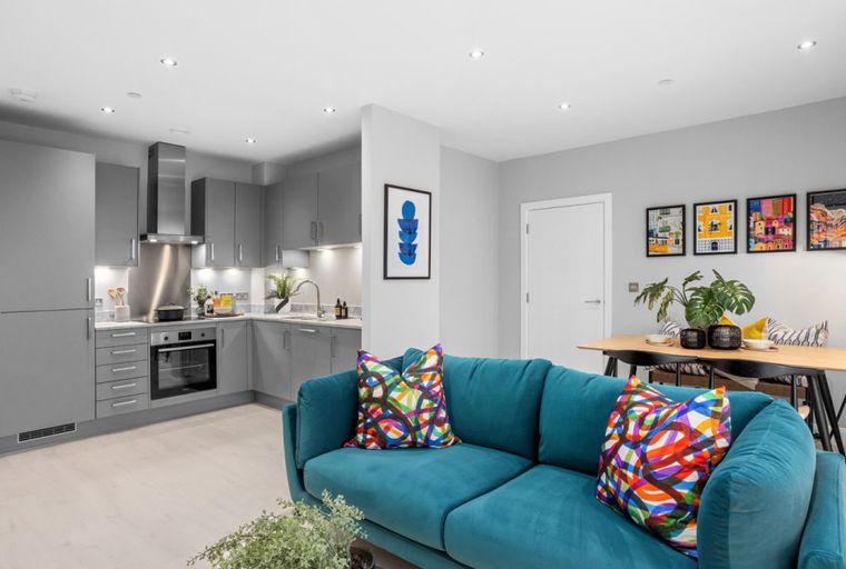 A living room and kitchen area with a teal sofa, dining table, and grey cabinets.