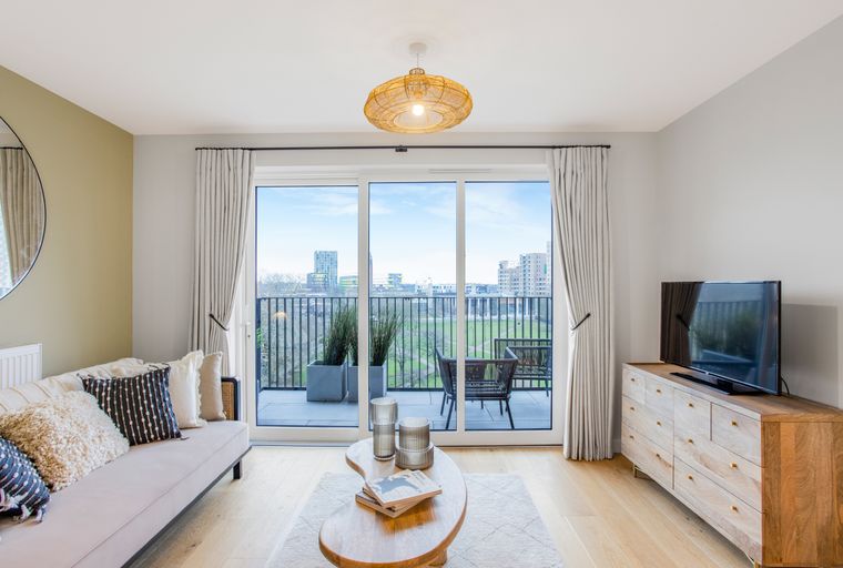 A living room with a sofa, coffee table, and a television, with a balcony visible through large glass doors.