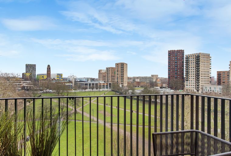 A view from a balcony overlooking a city park and modern buildings under a blue sky.
