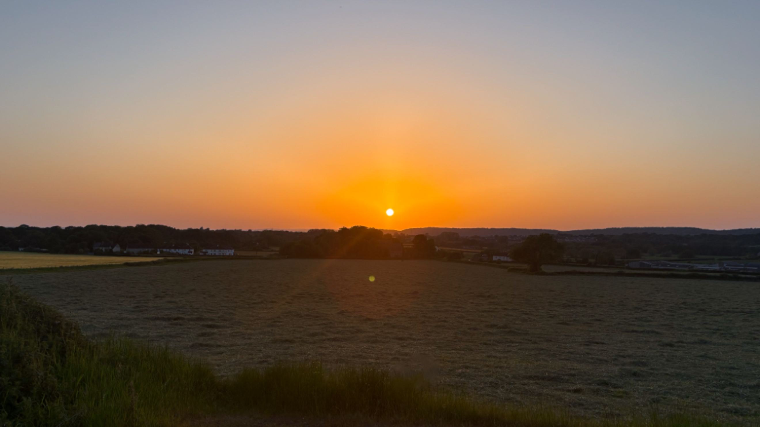 A sunset over a field with trees and houses in the background.