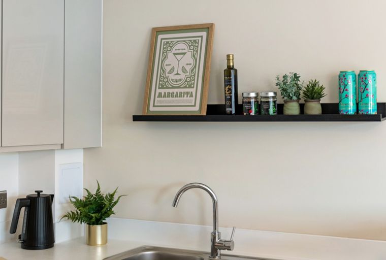 A kitchen counter with a sink, faucet, kettle, and a shelf with decorative items.