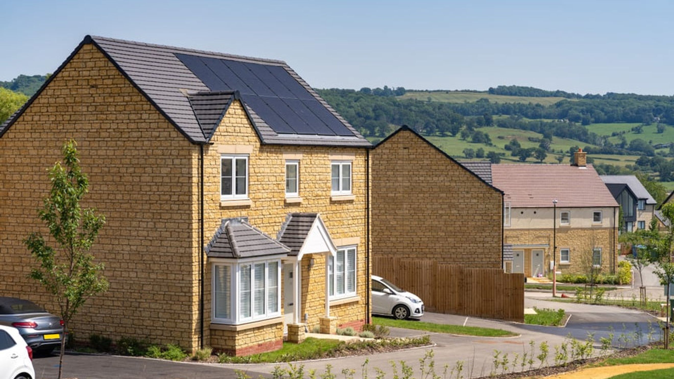 A row of modern houses with stone facades and solar panels on the roofs, set against a backdrop of green hills.