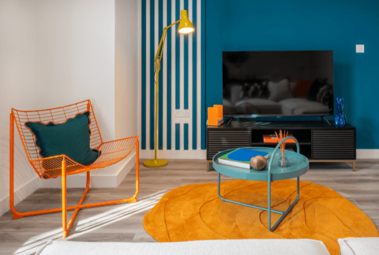 A living room with an orange chair, a yellow floor lamp, a television, and a teal coffee table on an orange rug.