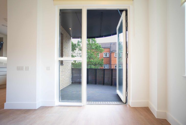 A view from inside a room looking out through open glass doors to a balcony with houses and trees visible.
