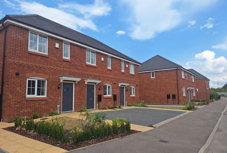 A row of new red brick townhouses with dark grey doors and white windows under a blue sky.