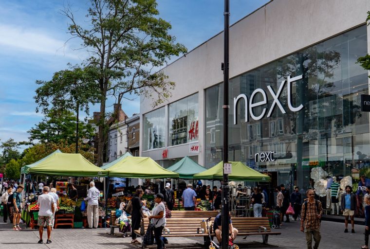 A busy street market with stalls and shops, including Next and H&M, with people shopping.